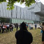 Crowd of students listening to a York University student (black female student in a black mid-length dress, long black hair in dreads), outside on a cloudy day. In front of the student building talking about student activism and resistance movements.