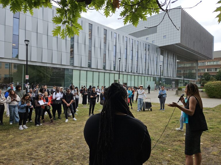 Crowd of students listening to a York University student (black female student in a black mid-length dress, long black hair in dreads), outside on a cloudy day. In front of the student building talking about student activism and resistance movements.